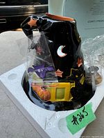 Glossy black ceramic witch hat spooky tealight house with moon and star cut-outs, Halloween themed decorations including a pumpkin, bat, and cat, sitting in original styrofoam packaging.
