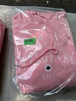 Photo of pink sweatshirt hoodie in sealed plastic bag showing front with white print text 'ORIGINAL LIFE' and maple leaf graphic.
