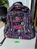 Front view of a black Roots backpack with colorful logos and text printed all over, showing zippered compartments and the Roots logo.