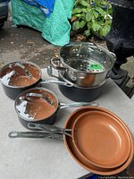 Set of Gotham Steel non-stick pots and pans showing two saucepans with copper-colored interiors and one larger pot with stainless steel steamer insert and glass lid, placed on a table outdoors.