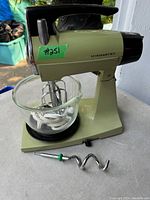 Front left view of vintage Sunbeam Mixmaster stand mixer with glass bowl and one metal attachment on beaters slot; another attachment lying in front of mixer