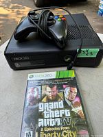 Black Xbox 360 console with wired controller on top and Grand Theft Auto IV game case in front.