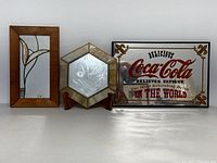 Three items displayed on a white surface: rectangular stained glass mirror with wooden frame on left, pentagonal stained glass mirror on wooden stand center, vintage Coca Cola mirrored bar sign on right.