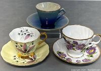 Three English style teacups with matching saucers displayed on table: yellow floral, white with purple flowers and blue cup with matching saucer
