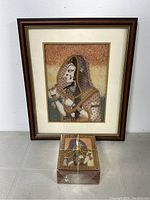 Framed Indian gem stone painting shown with small decorative box in front, both angled against a white wall on tiled floor.