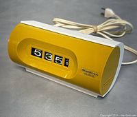 Side angled view of mustard yellow flip clock with black and white time display, power cord laid behind.