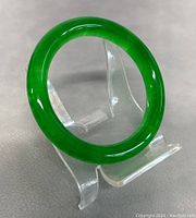Natural green jade bangle displayed on clear stand, showing smooth round shape and bright translucent green color.