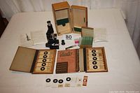 Overview photo of microscope, two wooden boxes with prepared slides, instruction manual, specimen tweezers, and additional glass slides arrayed on a white table.