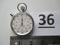 Full view of the silver-tone mechanical wind-up stopwatch showing dial, central red hand, outer black and red number markings, ring on top, and ruler showing 2 inch size.