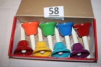 Photo of eight handbells arranged side-by-side in a box. Each bell is a different color: red, orange, yellow, green, light blue, blue, purple, dark red. White handles are visible attached to bells.