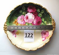 Top view of the large porcelain platter showing hand-painted pink and red roses with green and yellow foliage. Scalloped gold-trimmed edge visible. A ruler placed across the diameter shows size approximately 13 inches. Lot number tag 122 visible.