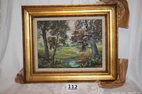 Oil painting in gold frame against white backdrop. Scene shows several trees flanking a creek with meadow background.