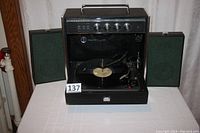 Front view of the Sylvania all-in-one record player with detachable speakers plugged in and stored behind the unit showing the turntable and control knobs.