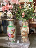 Two large Asian style ceramic vases side by side, the largest with floral motifs and pale green crackled glaze, the smaller with detailed painted figures and floral scenes, both with artificial flowers inside.