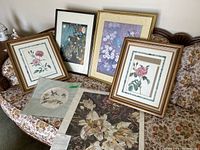 Wide view of five prints, three framed with floral themes, one figurative print framed, and two unframed prints laid out on sofa.