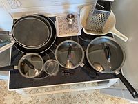 Photo showing three T-fal pots and frying pans with glass lids and black handles, a beige plastic strainer, granite-patterned trivet, and metal box grater on stovetop.