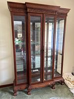 Front view of large wooden curio cabinet showing four glass paneled doors, carved decorative wood trim at crown and base, ball and claw legs, and internal glass shelves.