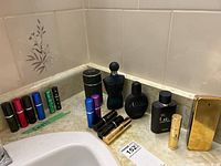 Photo showing full and travel-sized men's cologne bottles arranged on a countertop near a sink and tiled wall. Fragrances include Dark Obsession, 1 Million, Acqua Di Gio, Dunhill, and assorted small atomizers.