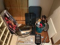 Photo of the entire kitchen lot showing a large stainless steel roasting pan with lid, wooden cutting boards, a teal utensil holder with various kitchen tools, a grill pan, a boxed cooking pan, a knife block, grater and meat tenderizer.