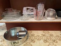 Full view of entire lot on two levels showing all items described including mixer, chopper, grinder, springform pan, measuring cup, sifter, and glass bakeware.