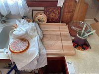 Image showing kitchen sink with various kitchen items arranged on the counter including a white oven mitt, a round trivet with a citrus fruit slice design, a wooden pastry board with engraved measurements, wooden cutting board, clear glass casserole dish with lid, vegetable patterned coaster, and a frying pan.