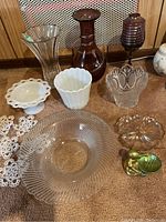 Overview showing multiple glass pieces, two milk glass items, a brown decanter, brass duck figurine, and crocheted doilies on carpeted floor