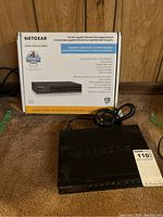 Front and boxed view of Netgear 24-port gigabit ethernet unmanaged switch with power cable coiled beside it on carpeted floor.