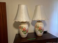 Pair of bedroom lamps with hand painted floral ceramic bases and matching cream shades placed on a wooden surface against a beige wall.