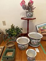 Full view of lot showing multiple Asian style pottery planters arranged on wooden floor next to a small wooden stand table. Some planters have artificial plants.