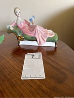 Photo showing front angled view of Royal Doulton figurine 'Repose' on a wooden table, depicting woman in pink dress reclining on chaise lounge holding a book.