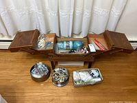 Full view of the wooden fold-out sewing basket with open compartments showing yarn, scissors, buttons, and other sewing notions inside and below the basket.