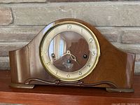 Front view of vintage wooden mantel clock on a wooden shelf with stone background, showing clock face and wooden casing with curved top.