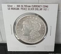 Obverse view of 1921 US Morgan Silver Dollar coin housed in a protective cardboard and plastic holder. Visible inscriptions include "E PLURIBUS UNUM" and year "1921". The coin shows lightly circulated condition.