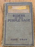 Front cover of the book showing title 'Riders of the Purple Sage' and author 'Zane Grey' with wear and damage visible on light blue cloth cover.