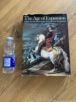 Photo of the large hardcover book The Age of Expansion next to a water bottle for size reference