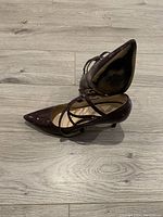 Side view of a dark burgundy patent leather pointed-toe heel with criss-cross straps, laid flat on wooden floor.