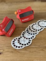 Two red vintage View-Master toys with several white circular reels laid out on a wooden surface, showing front view of toys.