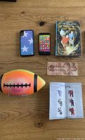 Photo showing all items in the lot: Hardy Boys book, rubber football, play cell phone, hockey postage stamps, and $2 bill spread out on a wooden table.