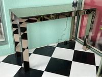 Front angled view showing full console table with smoked mirrored top and legs, in a black and white tiled floor background.