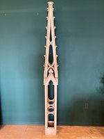 Full front view of the tall, thin, painted wooden obelisk against a blue-green wall, showing its entire height and base.