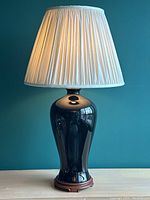 Full view of the lamp showing black ceramic body, pleated cream shade, and carved wood base on a wooden surface against teal wall.