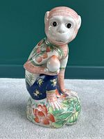 Front view of the hand painted Asian ceramic monkey figurine seated with a peach in its hand, detailed facial features, floral garment, and nature-inspired base.