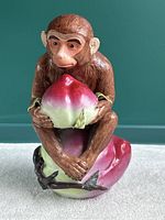 Front view of the hand painted ceramic monkey figurine sitting on a large fruit while holding a smaller fruit