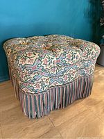 Side angle of the square ottoman showing tufted floral upholstery and multi-color fringe base
