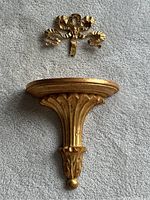 Top view of the gilt wall shelf with detailed carved design and gold finish next to the gold ribbon bow picture hanger.