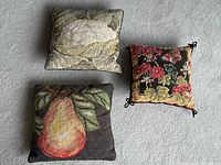 Three needlepoint accent pillows arranged on carpet showing different designs and sizes.