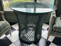 Front view of vintage round glass top table with green painted wicker base and visible black and white checkered floor beneath