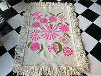 Full view of the rectangular cream wool rug with pink floral design and thick knotted fringe on all sides laid on black and white checkered floor.