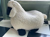 Side view of the sheep-shaped footstool showing its soft curly wool texture and black face and legs.