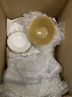 Top view of seven white plates stacked and one yellow bowl wrapped in bubble wrap inside a box.
